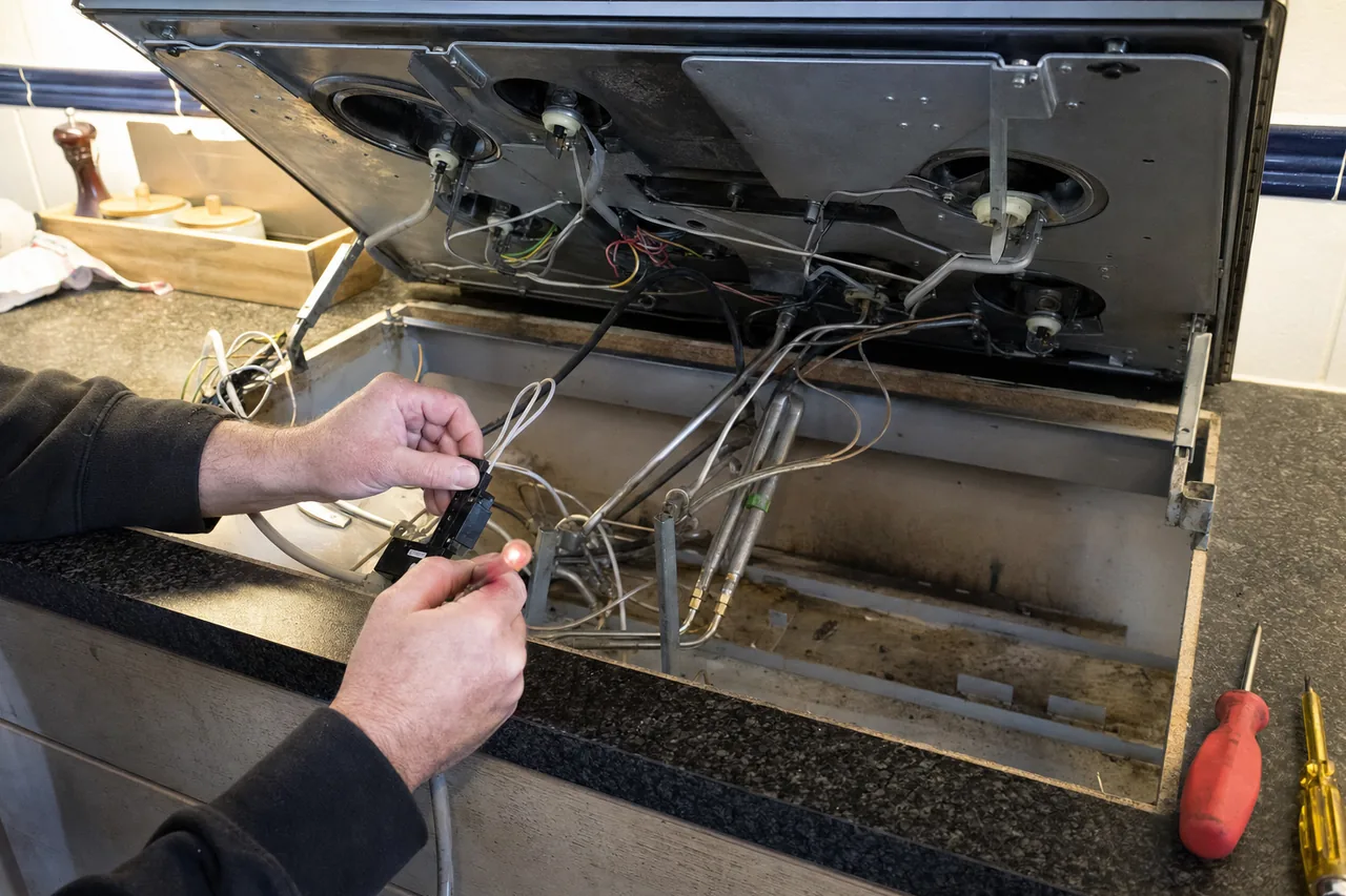 A hinged cooktop lid raised to show the burner underside, wiring loom and spark leads, with a spark module in hand